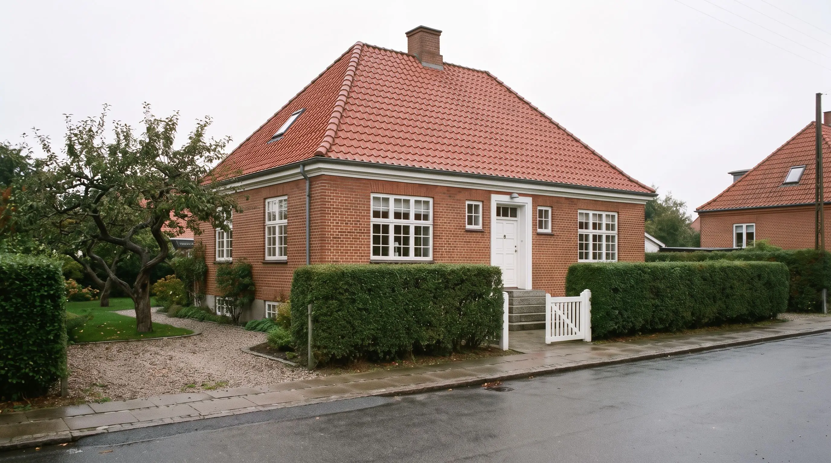 Murermestervilla i røde sten med hvide sprossede vinduer, rødt tegltag, klippet hæk og hvid havelåge i gråvejr