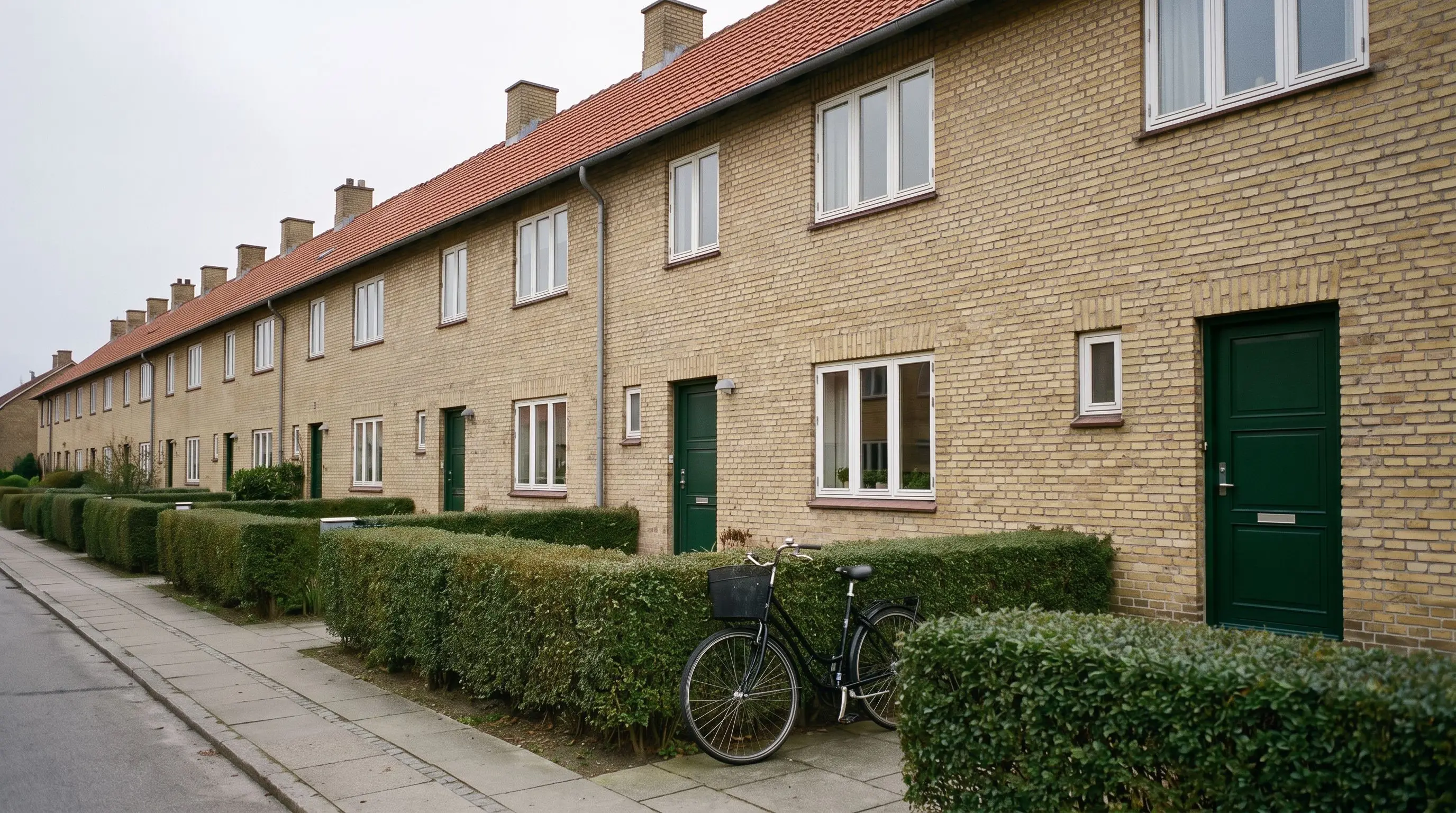 Række af gule murstensrækkehuse med rødt tegltag, hvide vinduer, mørkegrønne døre, klippede hække og en cykel ved fortovet