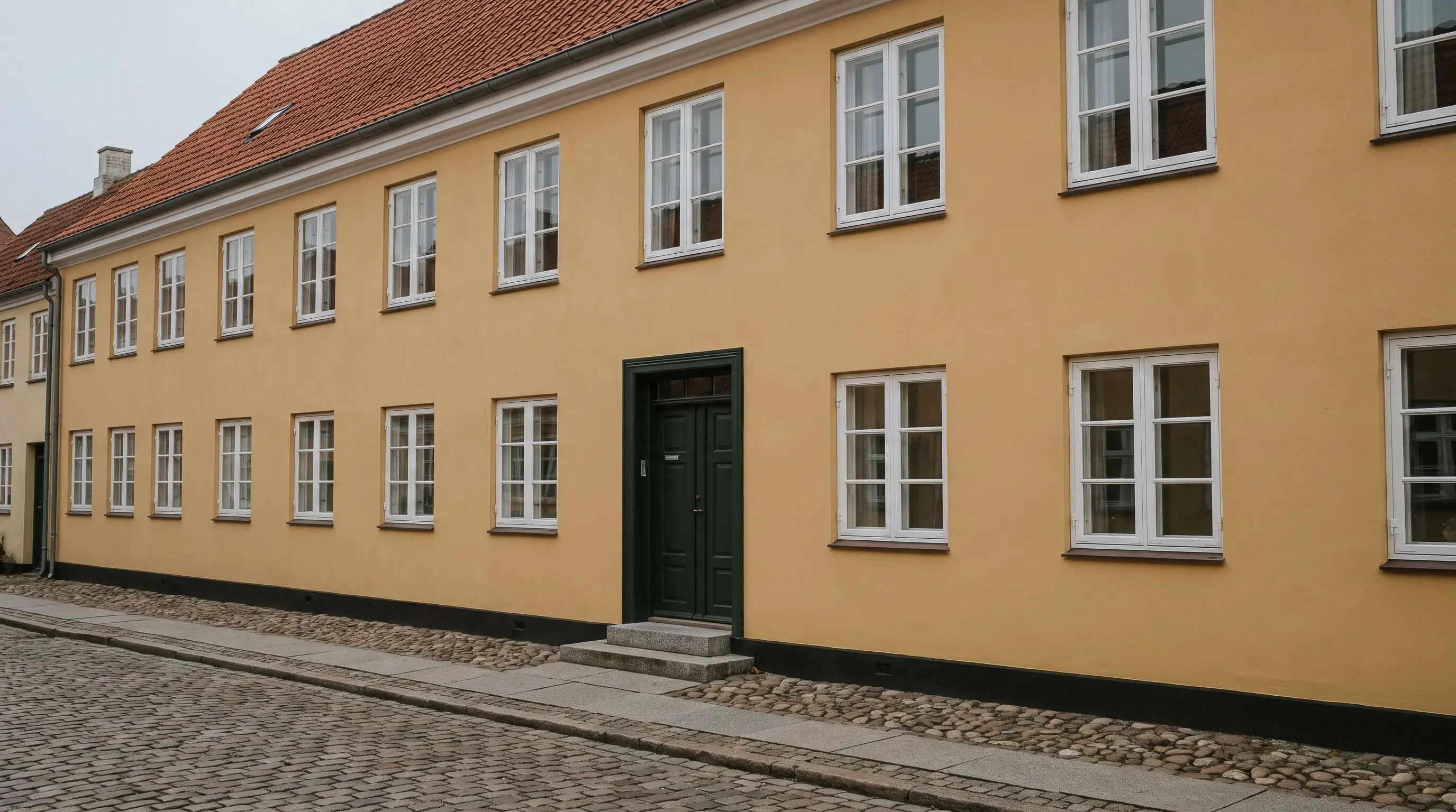 Ældre købstadshus i to etager med okkergul pudset facade, hvide sprossede vinduer, mørkegrøn fyldningsdør og brosten foran